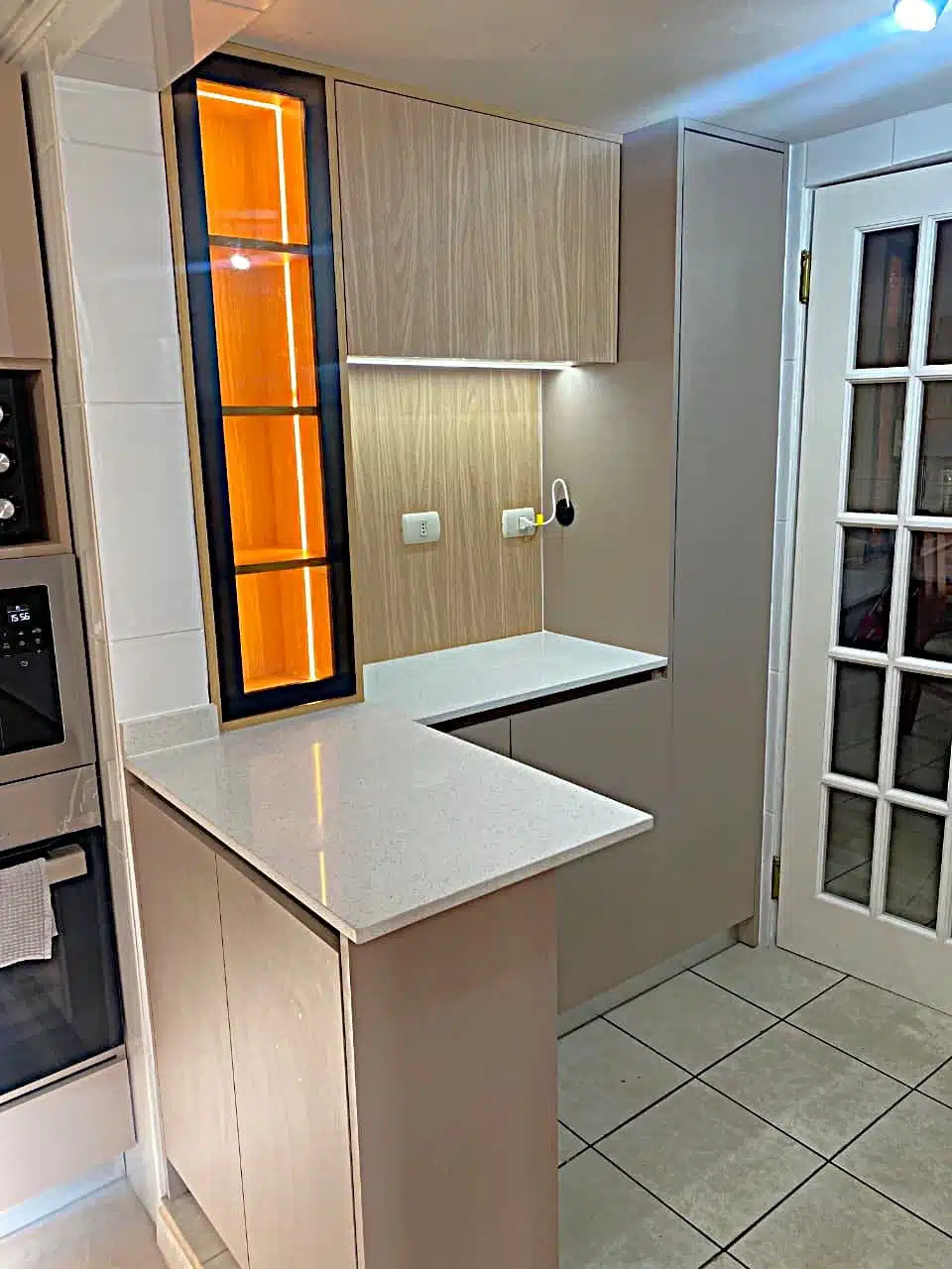 Muebles de cocina a medida en Santiago con cubierta de cuarzo, gabinetes en madera clara y vitrina vertical con iluminación LED.